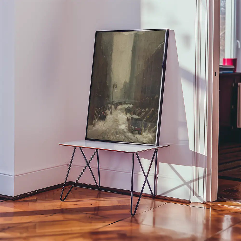 Framed painting depicting a winter street scene placed on a modern metal and wood table against a bright wall.