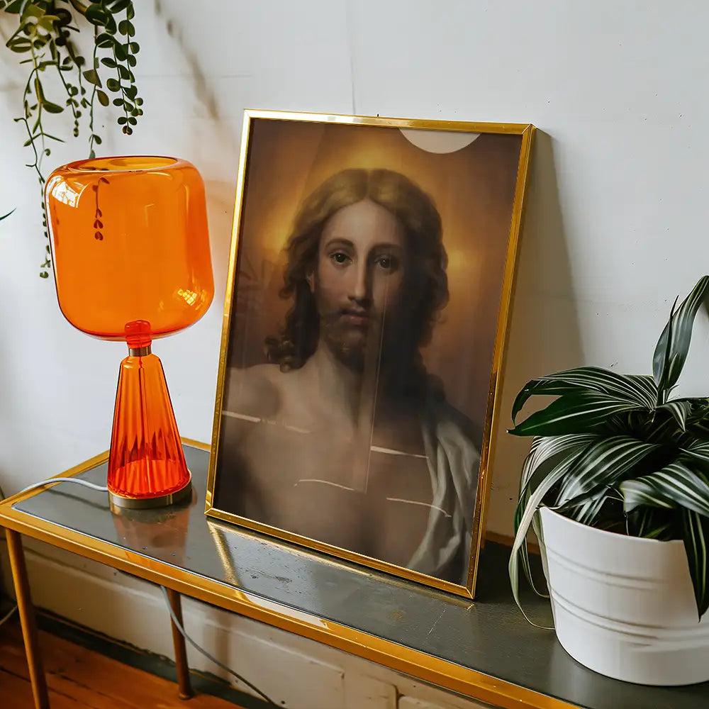 Framed painting of Jesus Christ displayed on a wooden console table with an orange lamp and a potted plant.