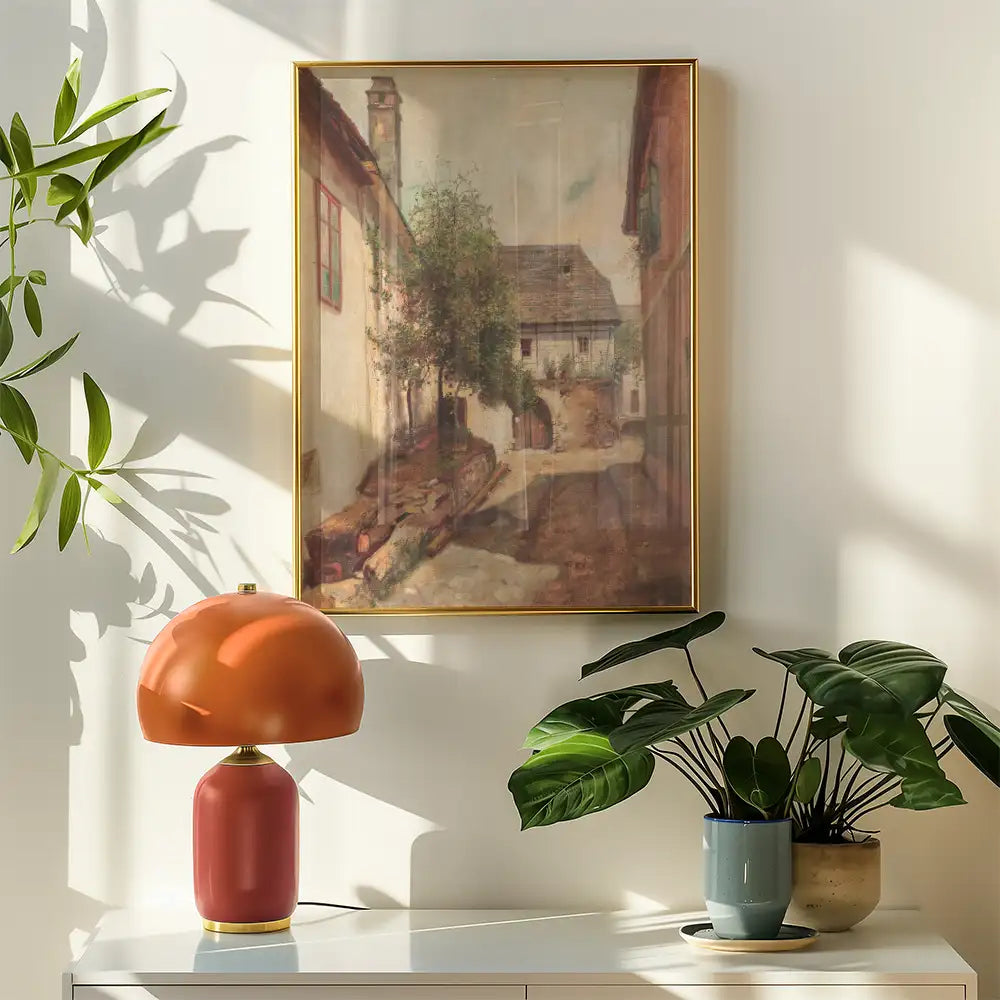 A vintage street scene painting with a golden frame, displayed on a wall beside a modern orange lamp and green potted plants.
