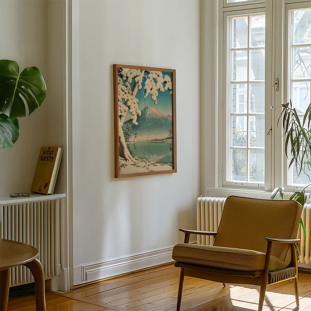 A framed painting of a snowy landscape with mountains and trees hanging on a white wall in a cozy living room with a plant and an armchair.