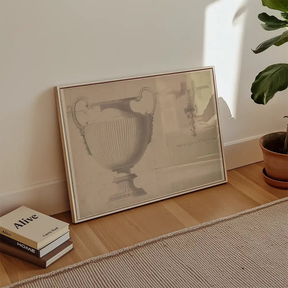 Framed artwork depicting a classical urn design, leaning against a wall with decorative plants and books in the foreground.