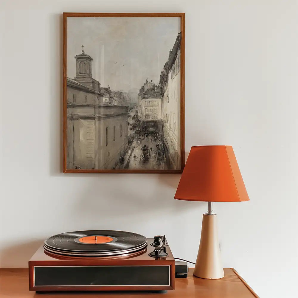 Framed painting of a street scene displayed above a vintage record player and an orange lamp on a wooden table.