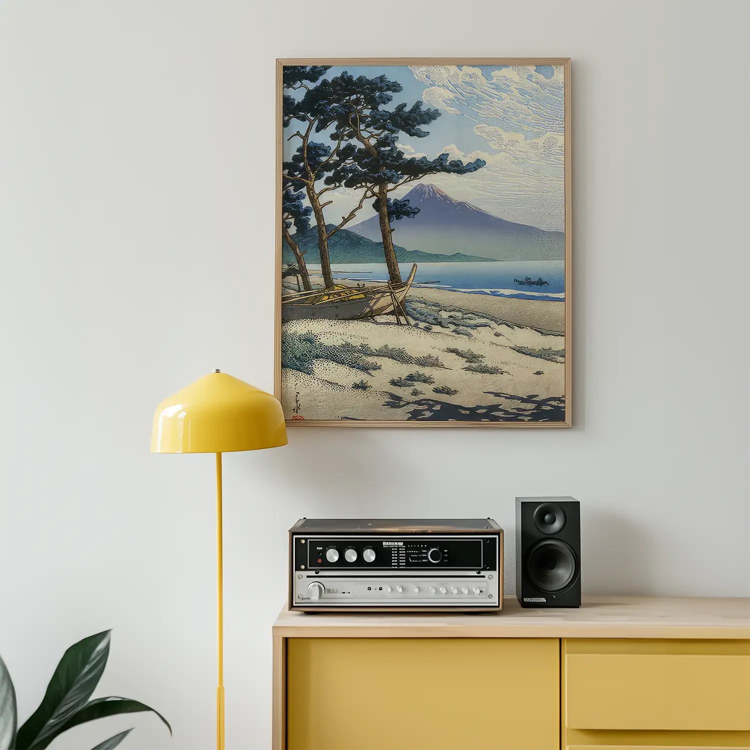 Framed painting of a serene beach scene with trees and mountains, displayed above a yellow lamp and audio equipment on a wooden cabinet.