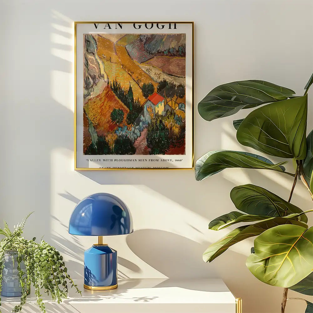 Framed painting of "A Valley with Ploughman Seen from Above, 1889" by Van Gogh, displayed on a wall with a blue lamp and green plant.