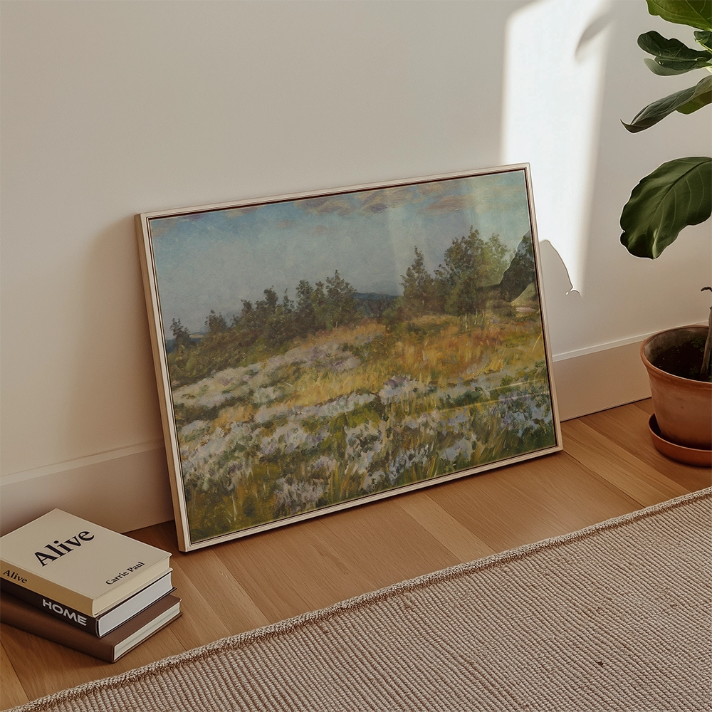Framed landscape painting depicting a vibrant field with trees in the background, placed next to a stack of books and a potted plant.