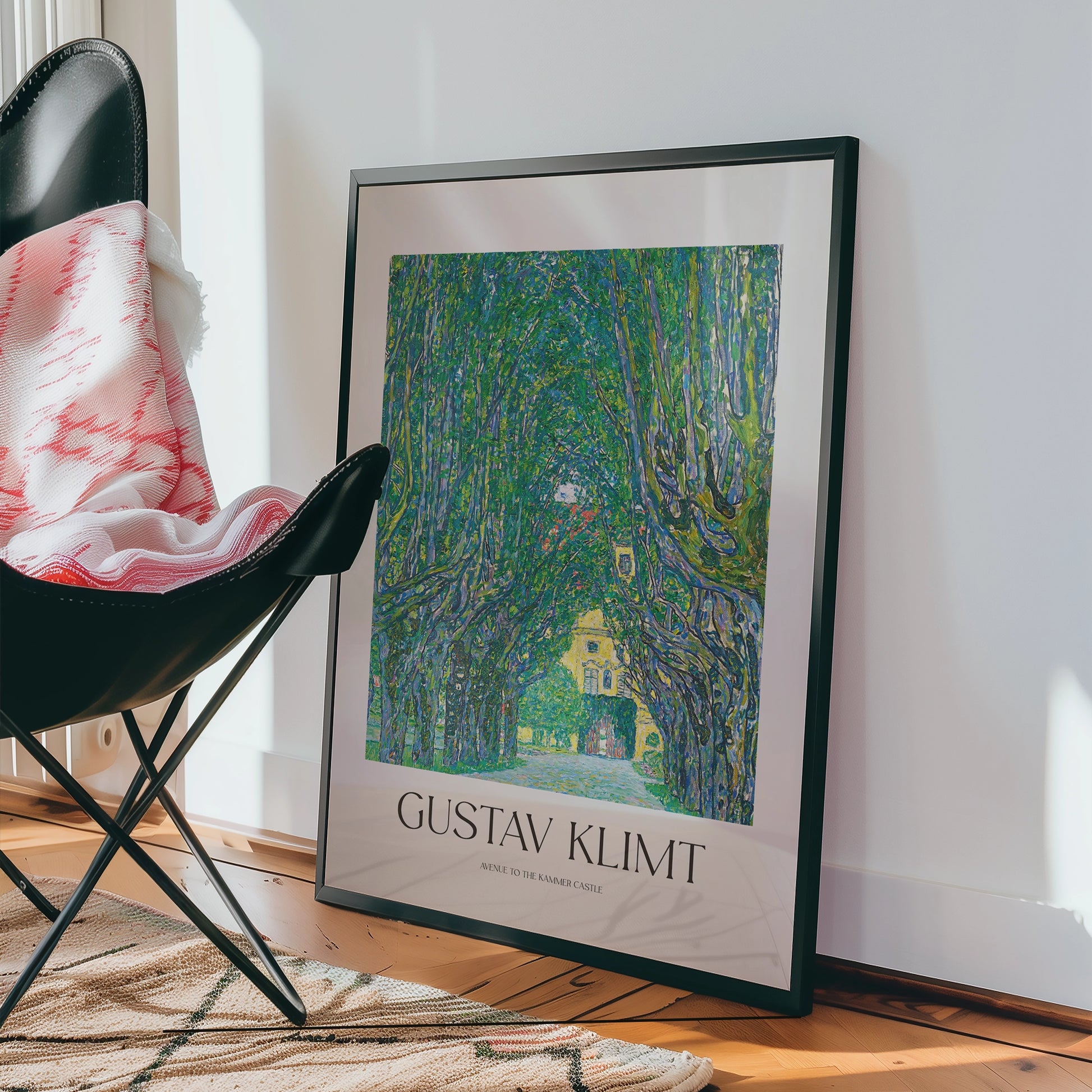 Gustav Klimt poster featuring a vibrant green tree-lined path leading to a yellow building, elegantly framed and displayed beside a black chair with a patterned throw.