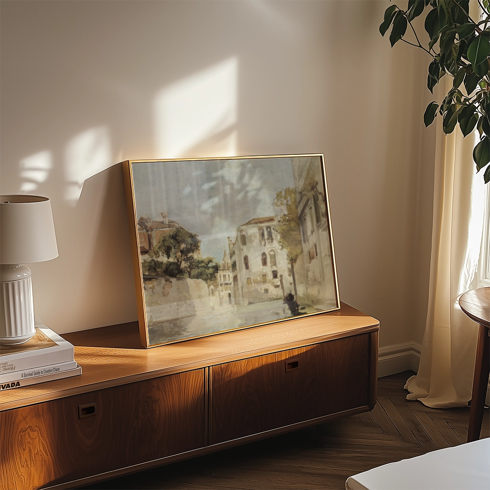Painting displayed in a modern living room setting, framed in gold, depicting a serene street scene with buildings and trees.