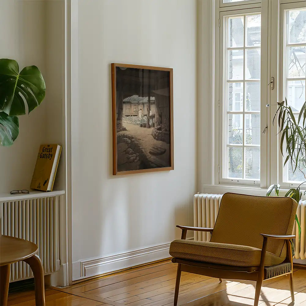A framed painting depicting a serene outdoor scene with a rustic cottage and figures, displayed in a modern interior with natural light and indoor plants.