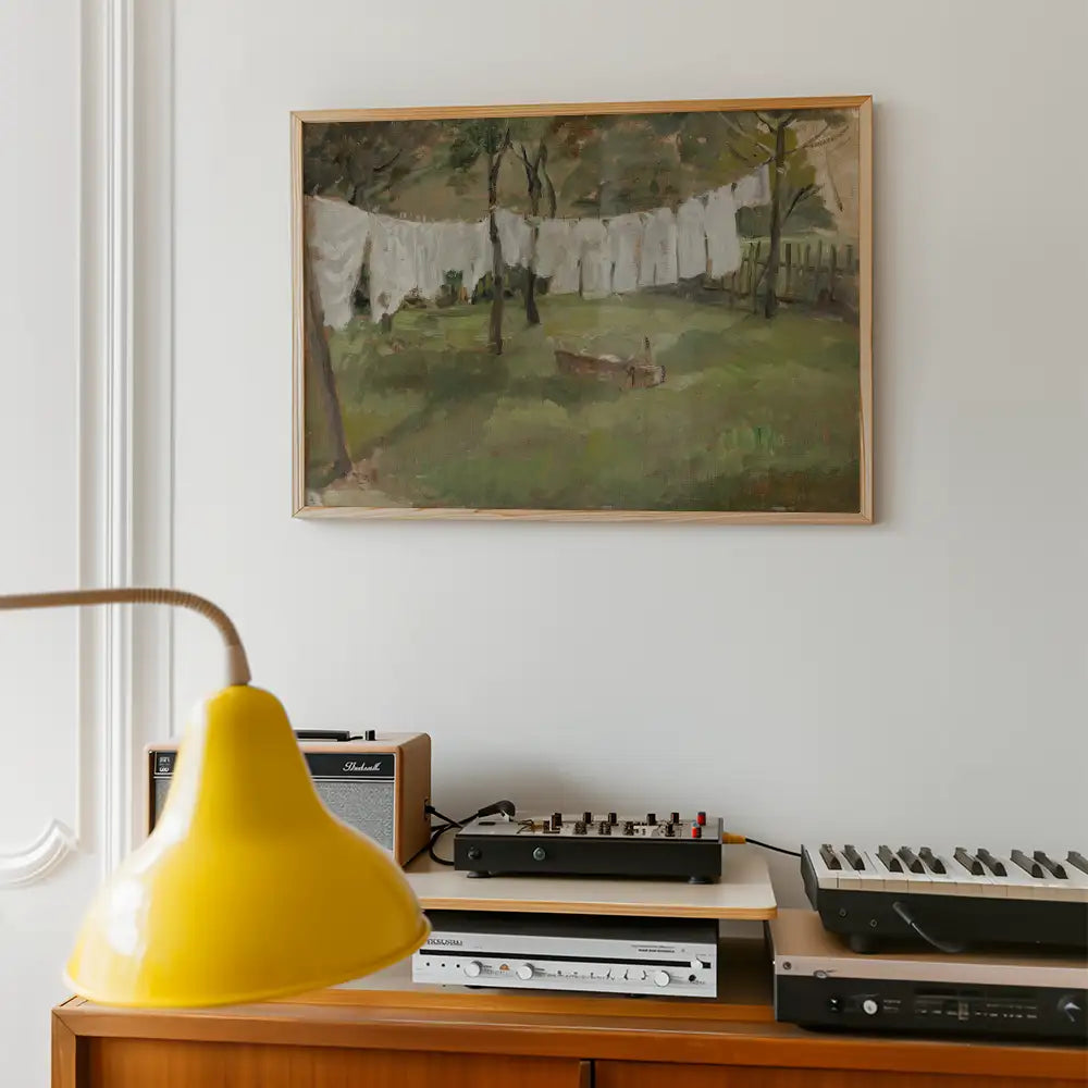 Painting of a serene outdoor scene with laundry hanging on a line in a green garden, framed and displayed on a light-colored wall, alongside a yellow lamp and vintage audio equipment.