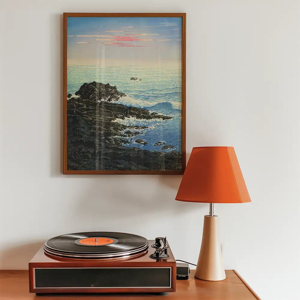 Painting of a seascape with rocks and waves, framed and displayed above a vintage record player and an orange lamp.
