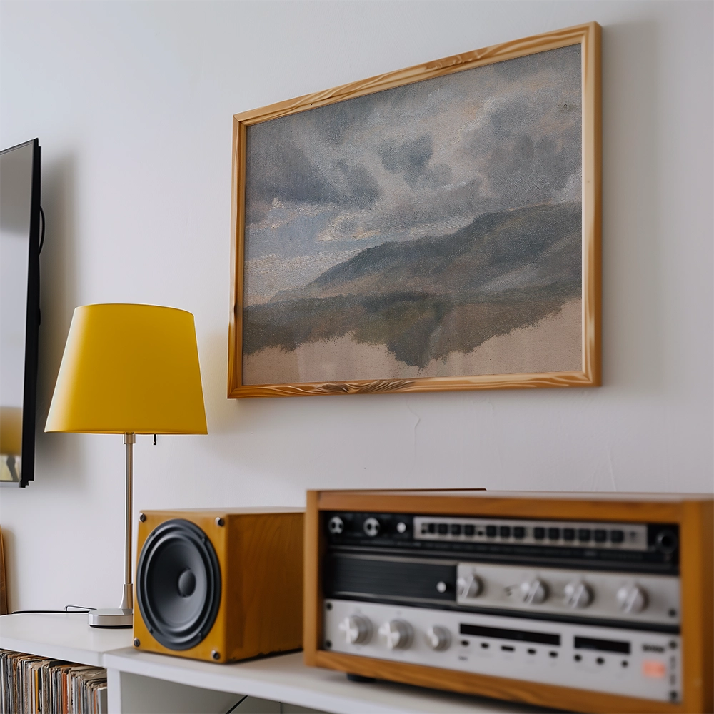 Painting of a serene landscape with mountains and clouds, framed in natural wood, displayed on a wall beside a yellow lamp and vintage audio equipment.