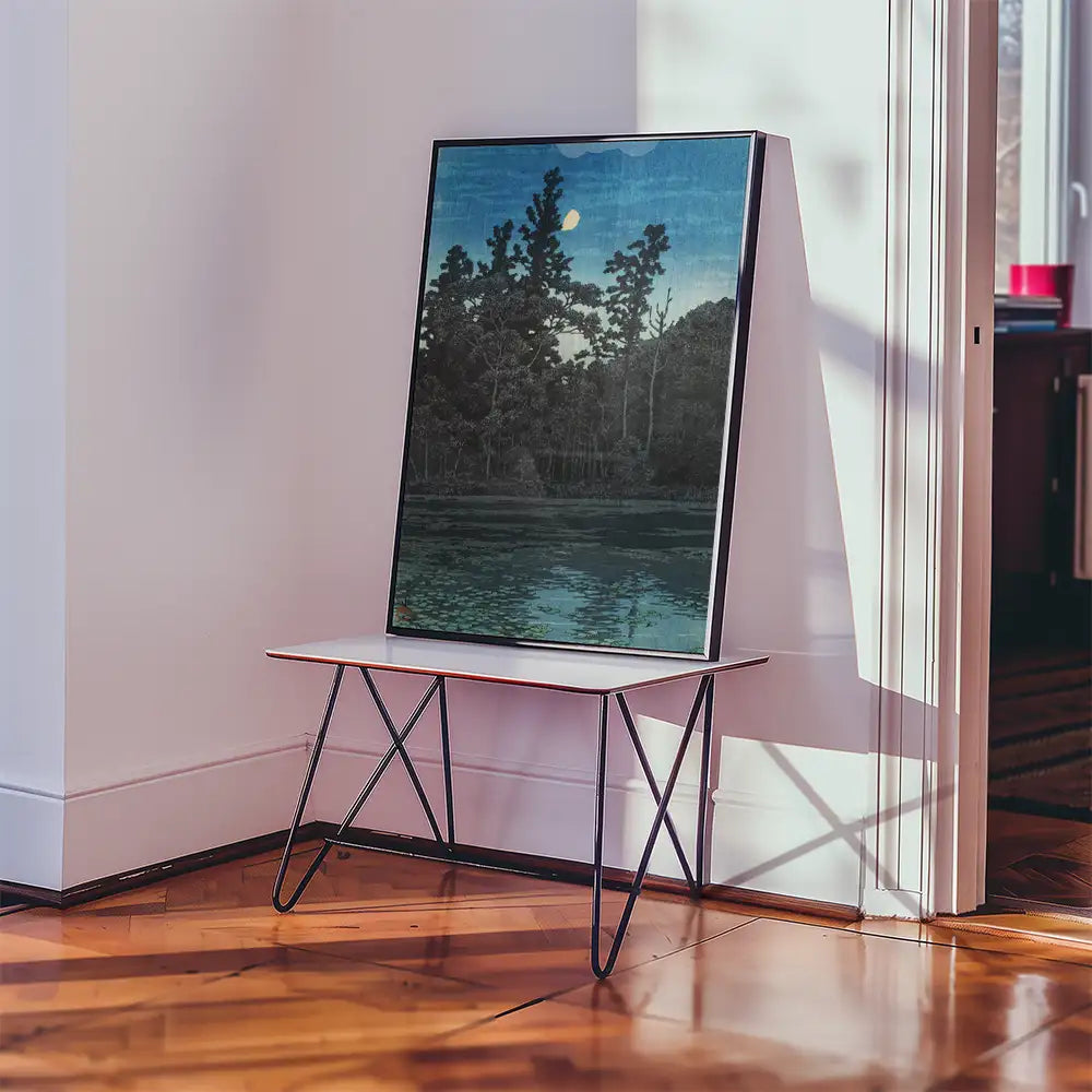 A framed landscape painting of trees by a still body of water displayed on a minimalist table in a well-lit room.