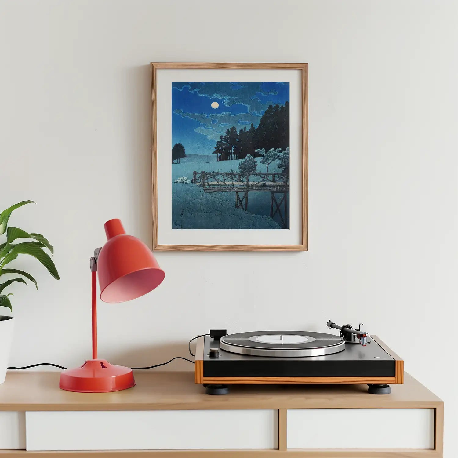 Framed painting of a snowy landscape with a moonlit sky, displayed above a vintage record player and a red table lamp on a wooden surface.