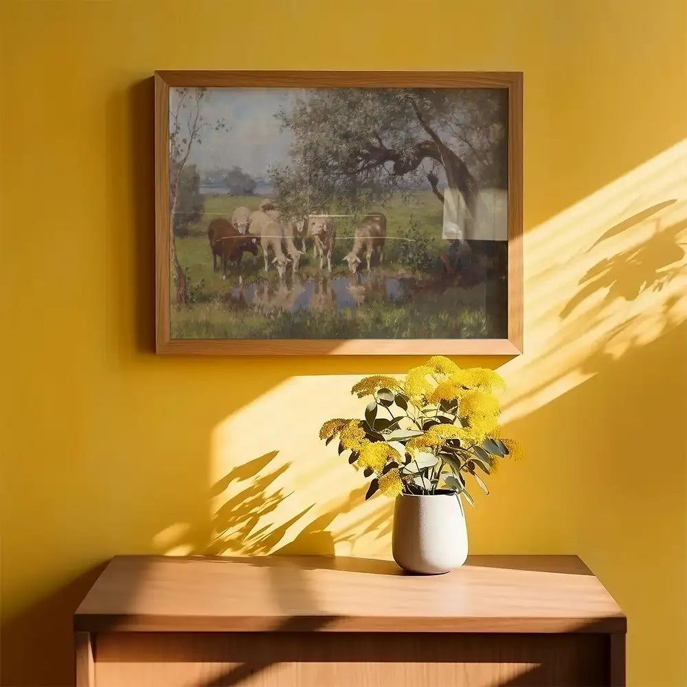 Pastoral painting of cows by a water body in a wooden frame, hung on a vibrant yellow wall, with a vase of yellow flowers on a wooden surface below.