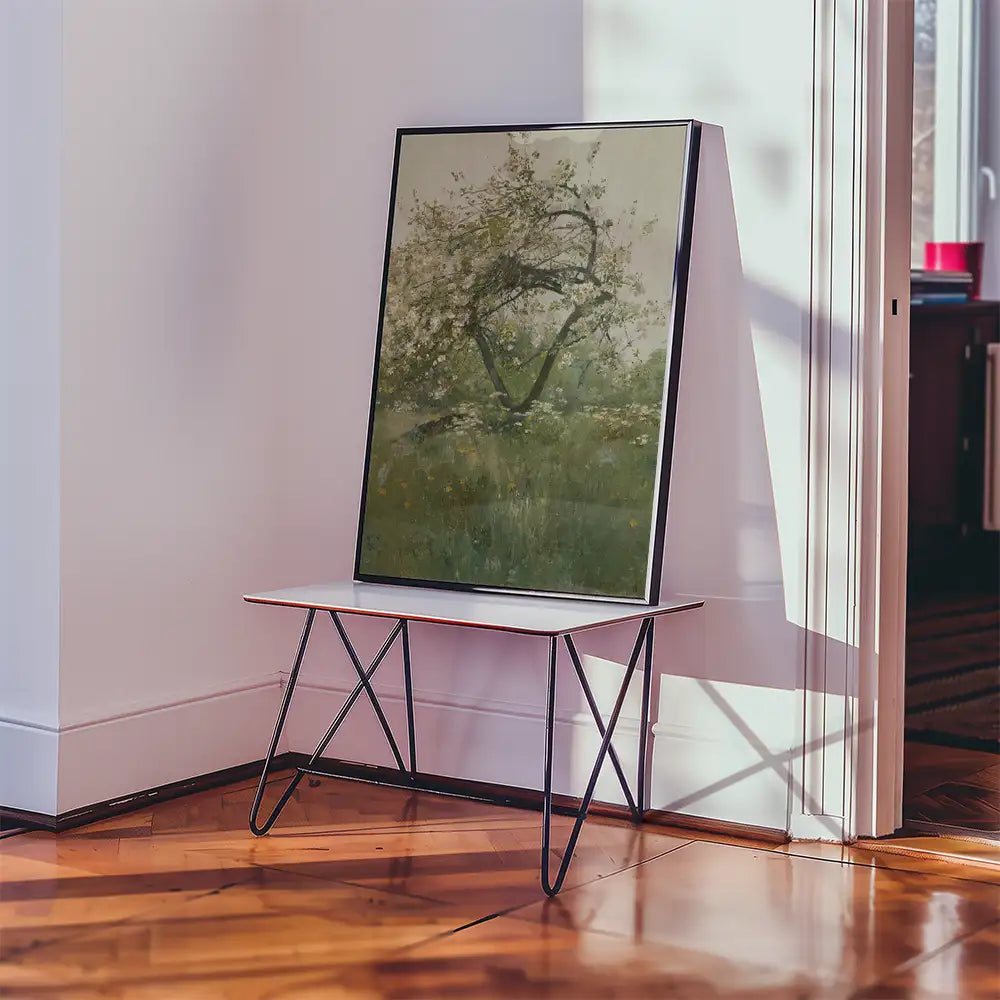 Framed painting of a tree in a grassy field displayed on a modern table in a bright interior setting.