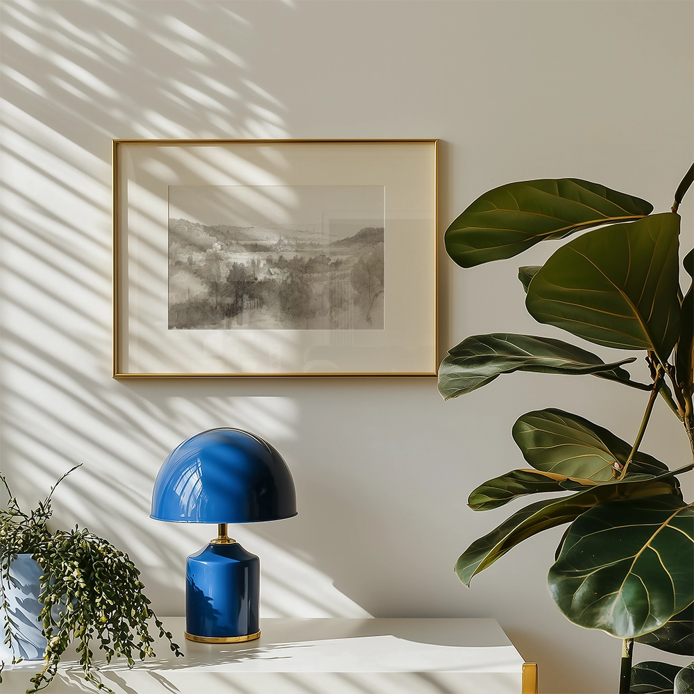 Framed landscape painting in soft greys and browns, displayed on a wall with a blue lamp and green plant.