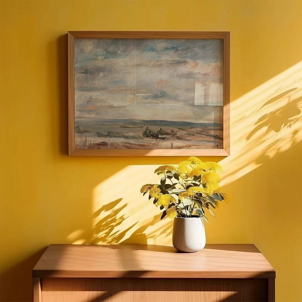 Framed landscape painting on a yellow wall with a vase of yellow flowers on a wooden table.