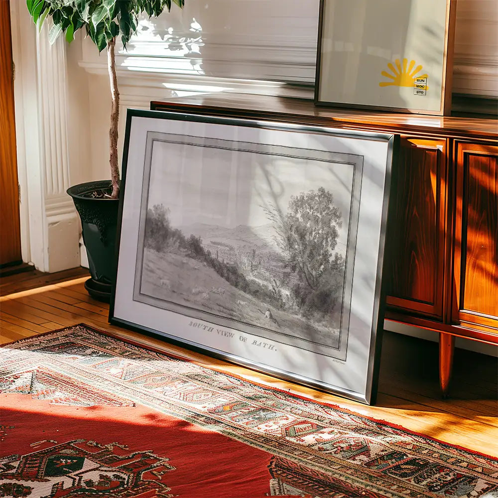 Framed black-and-white landscape painting titled "South View of Bath," leaning against a mid-century modern wooden sideboard with a potted plant and traditional rug in a sunlit room.