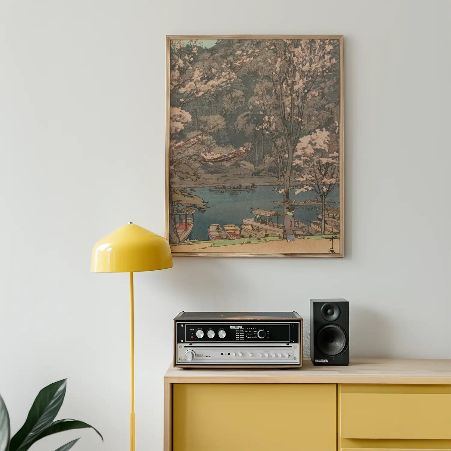 Framed painting of a serene landscape with cherry blossom trees and boats by a river, displayed on a light wall next to a yellow lamp and a stereo on a wooden cabinet.