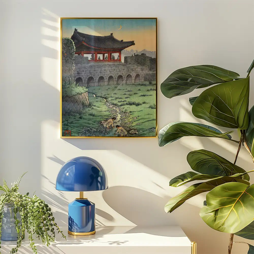 A framed painting of a traditional landscape featuring a pagoda and figures in a lush green setting, displayed on a wall beside a blue lamp and a potted plant.