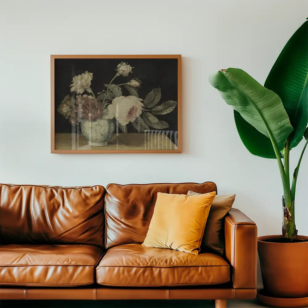 A beautiful floral painting in a wooden frame displayed above a brown leather couch with a decorative yellow pillow and a green plant beside it.
