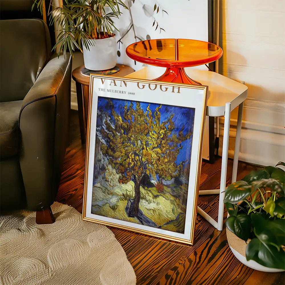 Framed painting of "The Mulberry" by Vincent van Gogh, featuring a vibrant tree against a blue background, displayed in a cozy living space with a plant and modern furniture.