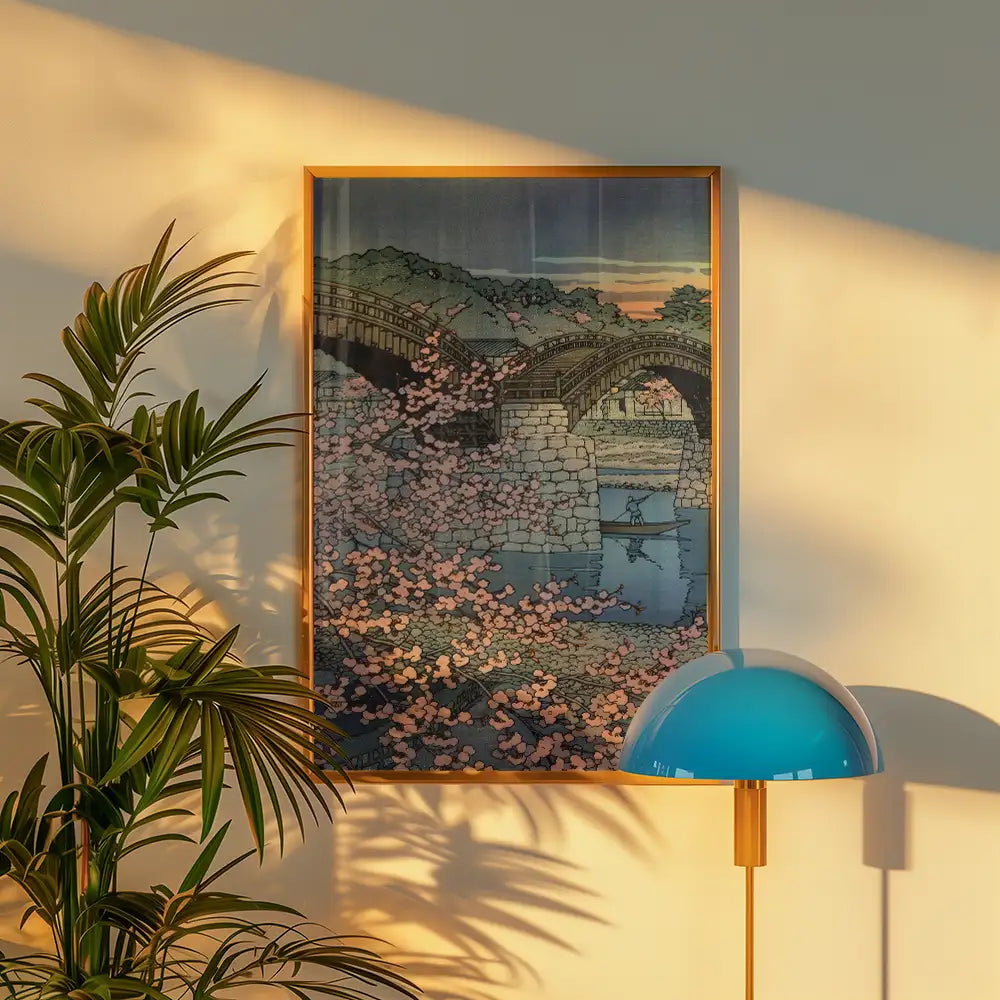 Framed painting of a bridge surrounded by cherry blossoms, displayed on a wall next to a plant and a modern lamp.