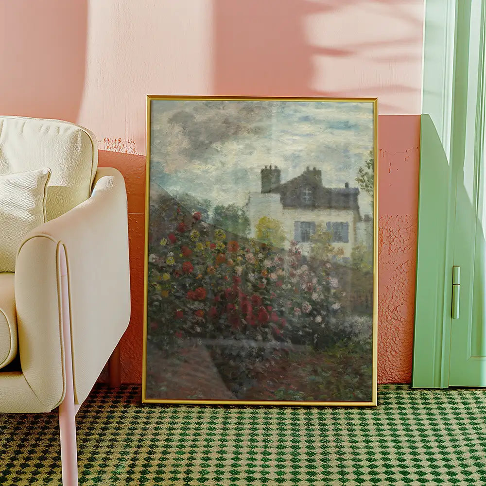 Framed painting of a house surrounded by blooming flowers, leaning against a light-colored armchair in a stylish interior setting.