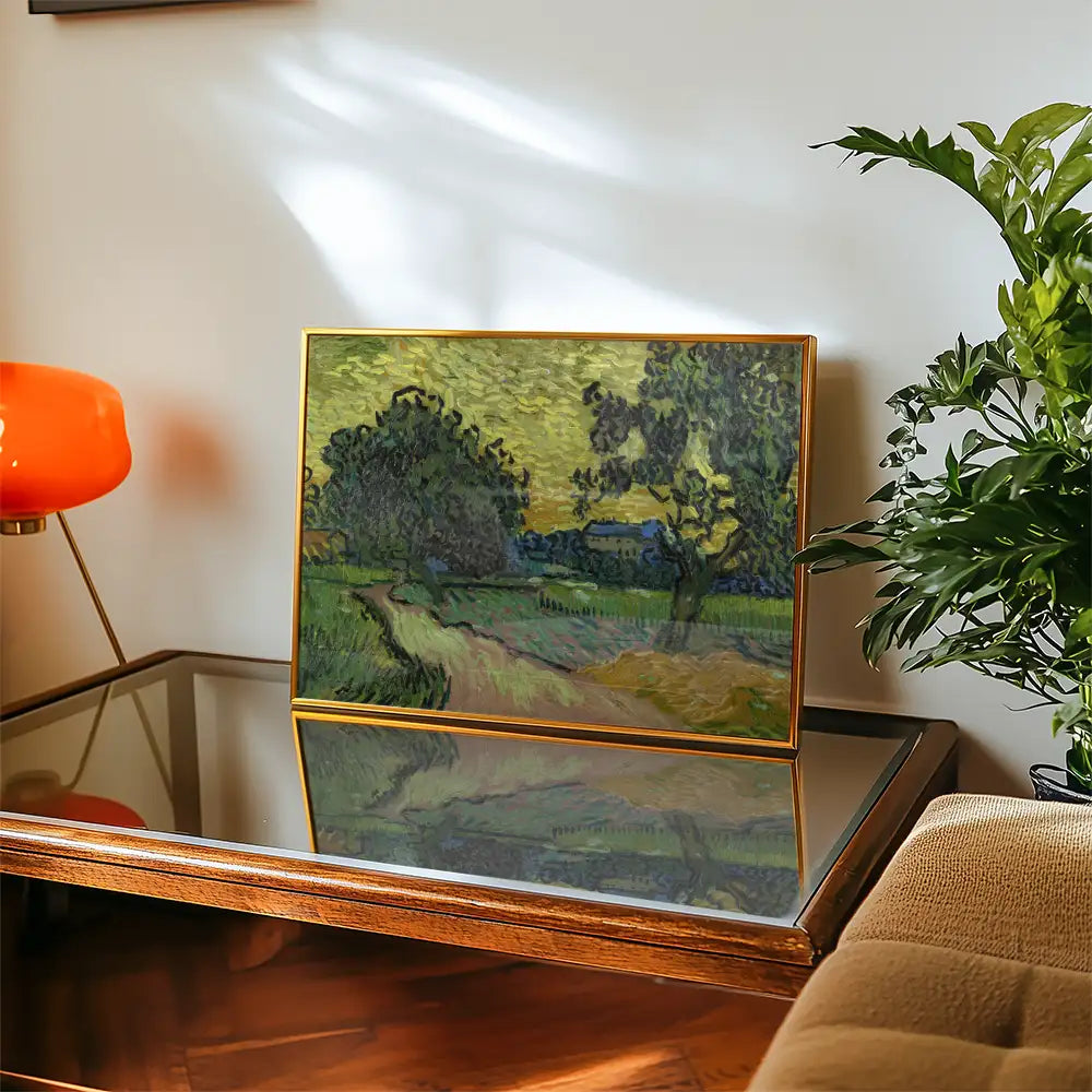 Framed landscape painting featuring trees and a path, displayed on a wooden table alongside a modern lamp and plant.