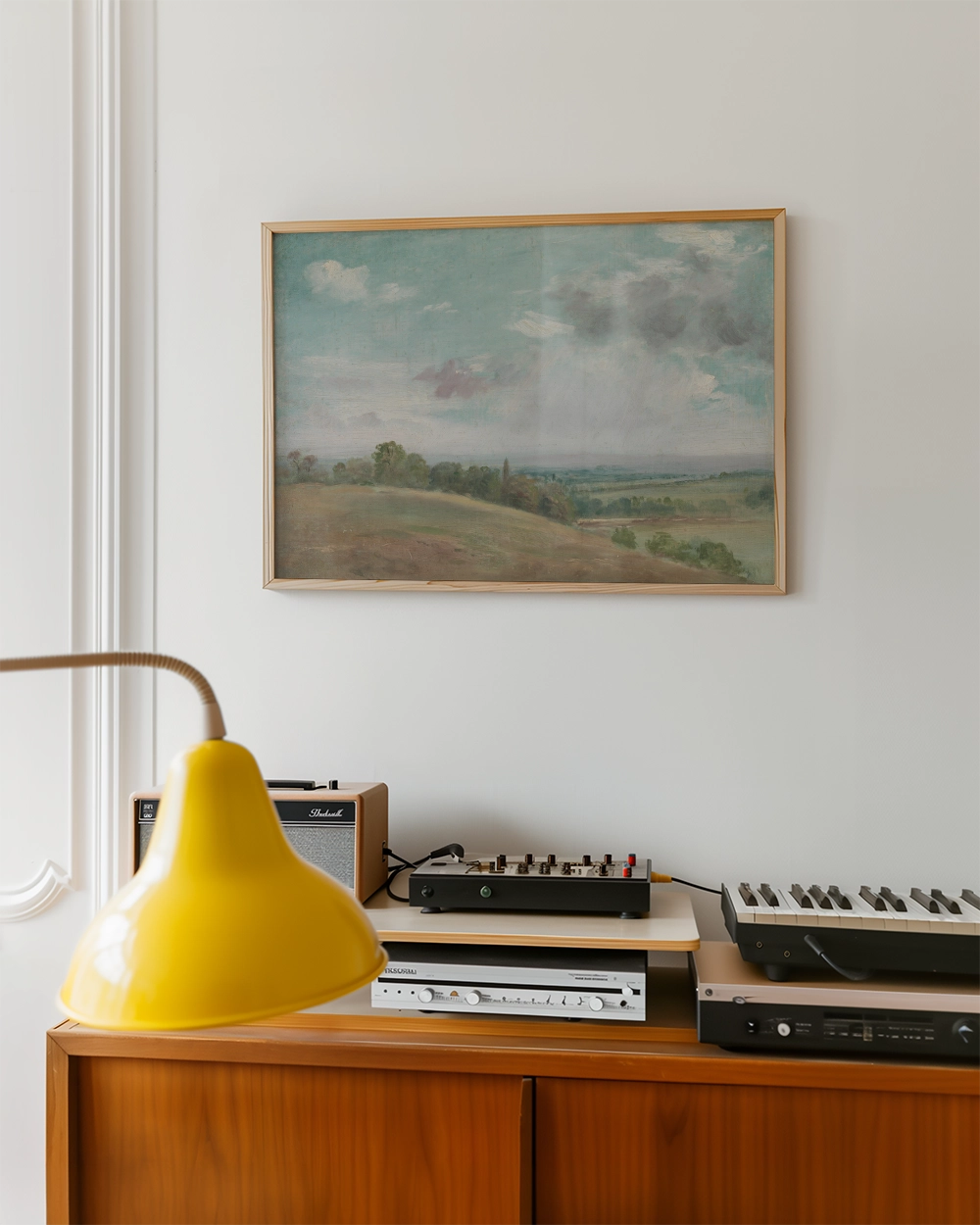 Framed landscape painting hanging on a wall above a wooden cabinet with a yellow lamp and audio equipment.