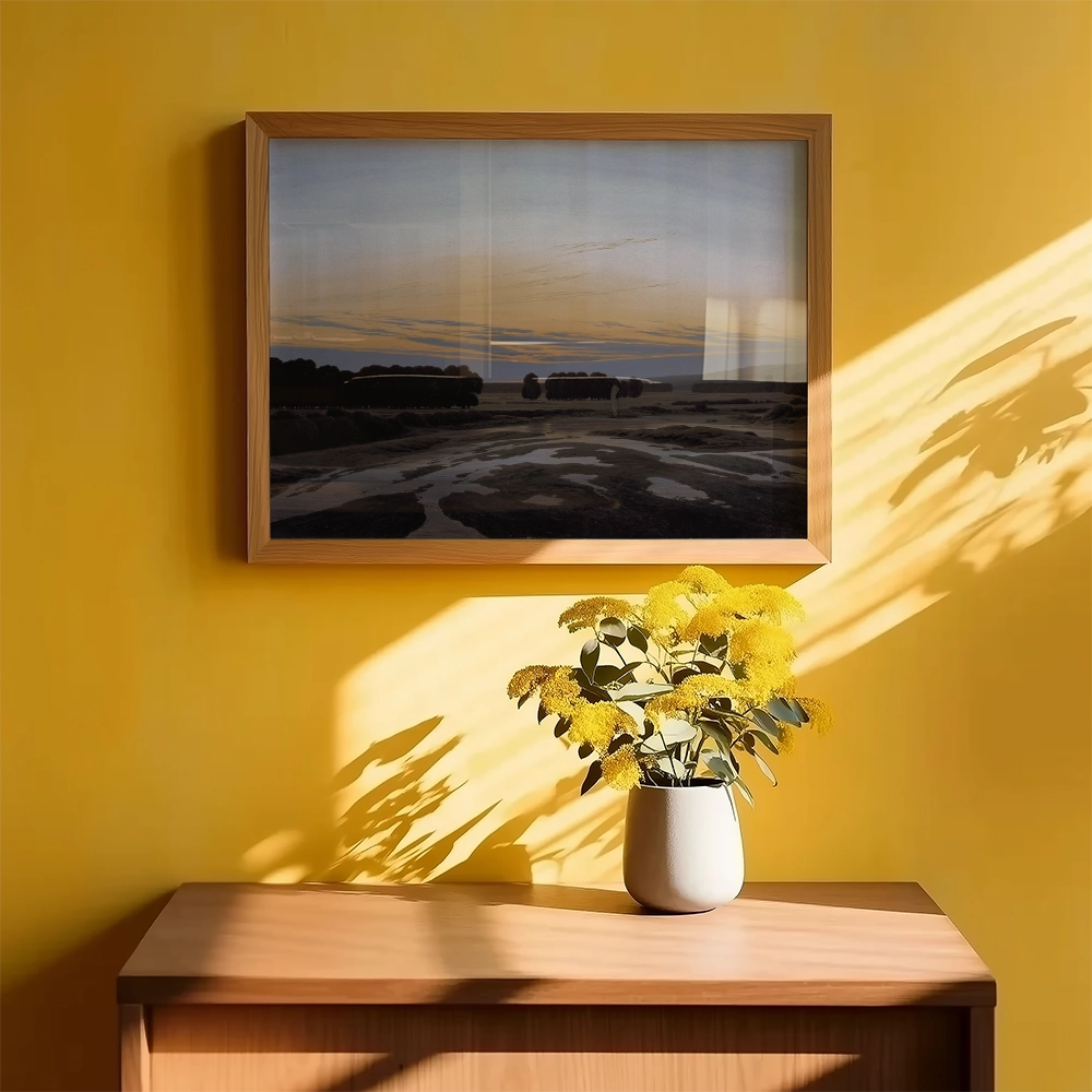 Framed landscape painting hanging on a yellow wall above a wooden cabinet with a vase of yellow flowers.