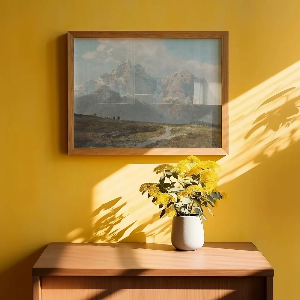 Framed mountain landscape painting above a wooden console table with a white vase of yellow flowers, set against a bright yellow wall.