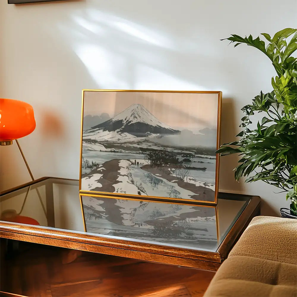 Framed painting of a mountain landscape displayed on a wooden table next to a plant and an orange lamp.