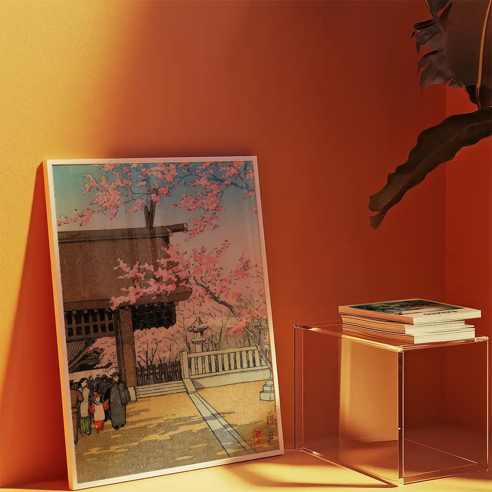Framed painting of a cherry blossom scene with people walking in front of a traditional structure, set against an orange wall with a clear display table holding stacked books.