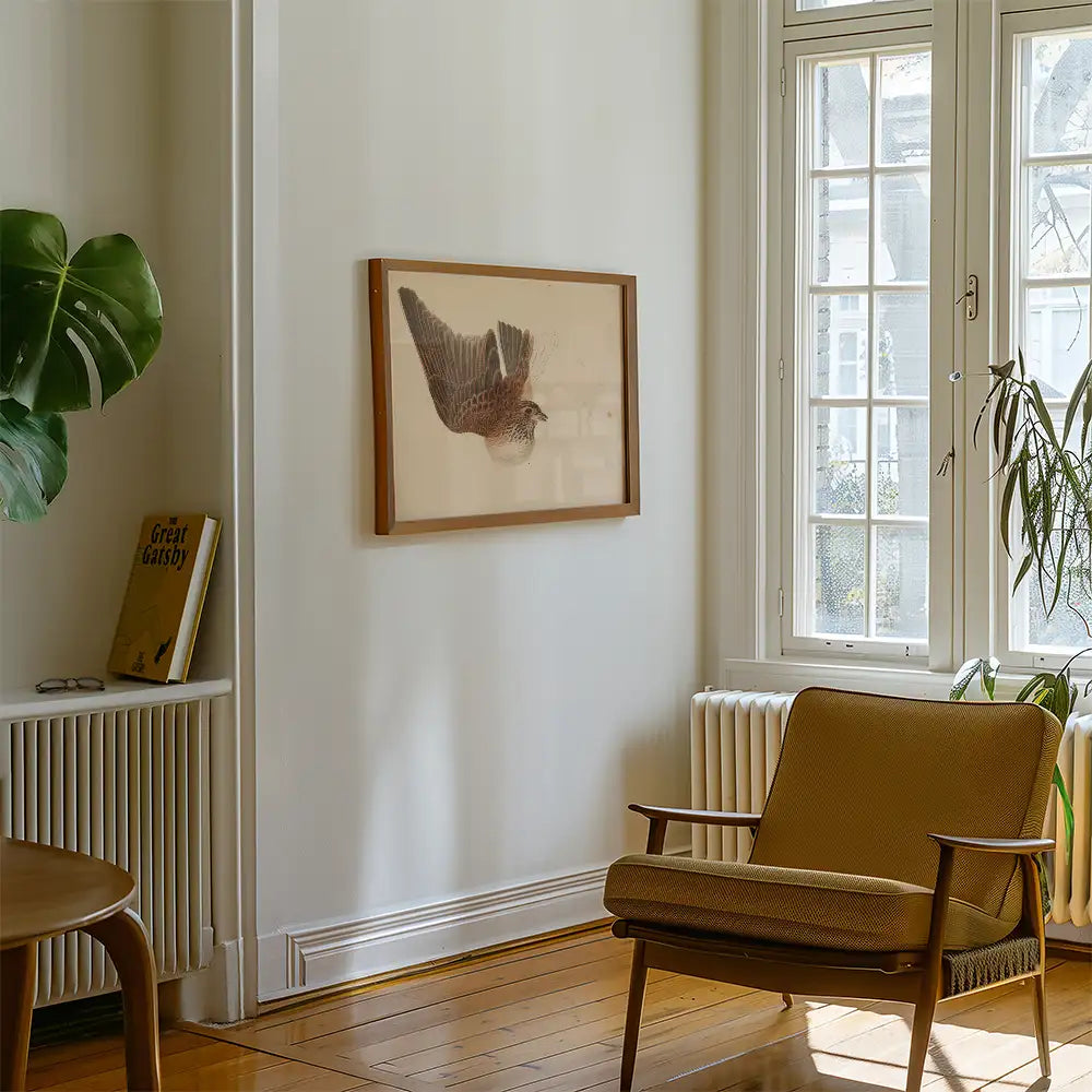 Framed artwork of a bird displayed on a wall in a cozy living room setting with a plant and mid-century chair.