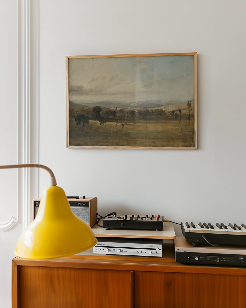 Framed landscape painting hanging on a white wall above a wooden console with a yellow lamp and various audio equipment.