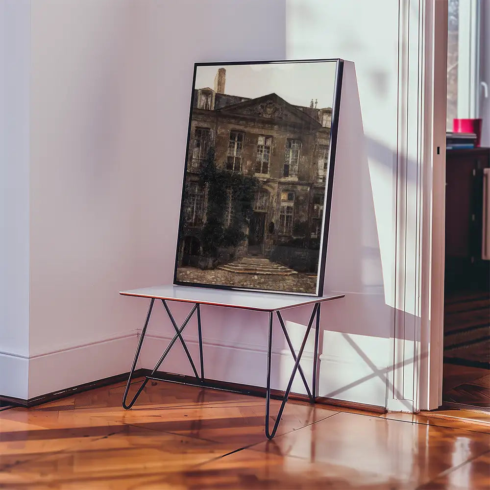 Framed painting of an old building with a stone pathway displayed on a minimalistic table in a bright interior setting.