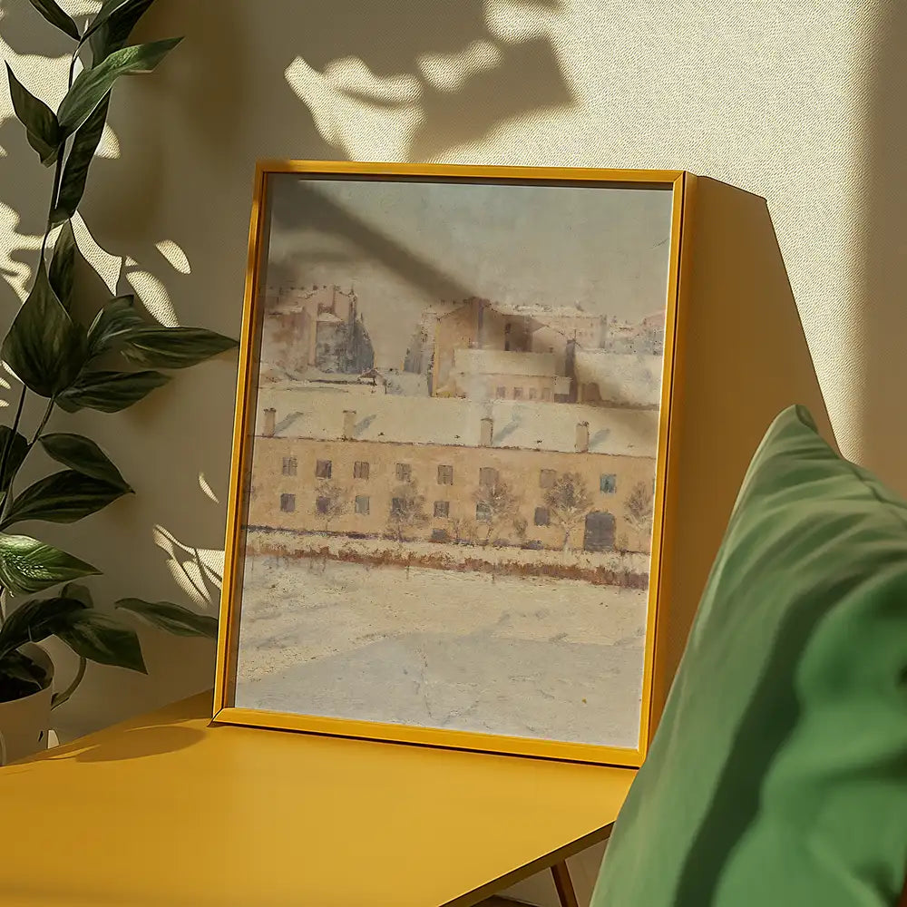 Painting in a yellow frame depicting a winter landscape with buildings, placed on a yellow table beside a green cushion and a plant.