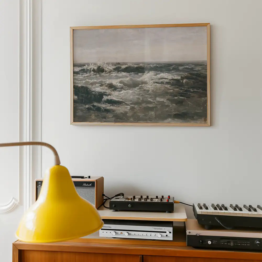 Framed painting of ocean waves hanging on a white wall, with a yellow lamp and music equipment in the foreground.