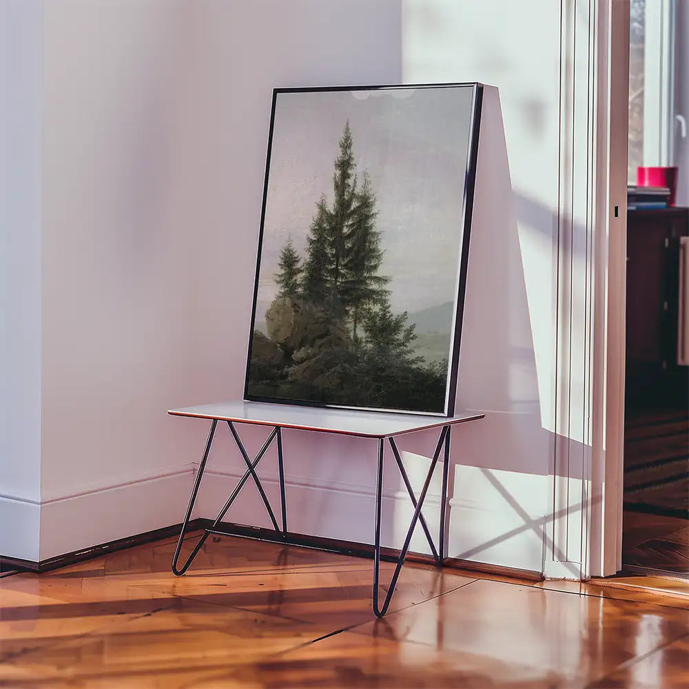 Framed painting of pine trees and mountains displayed on a modern white table in a well-lit interior setting.