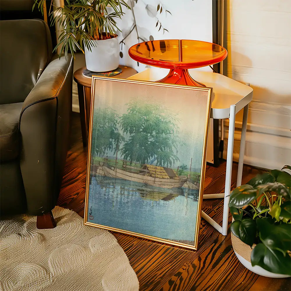 Framed painting of a serene riverside scene featuring a boat, surrounded by trees, displayed in a cozy living room setting.