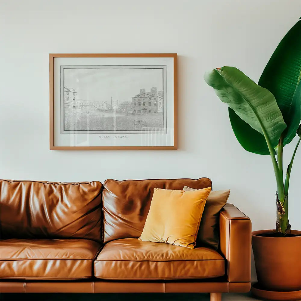 Framed black and white painting of a historical square displayed on a wall above a brown leather sofa with a yellow cushion and a large green plant.