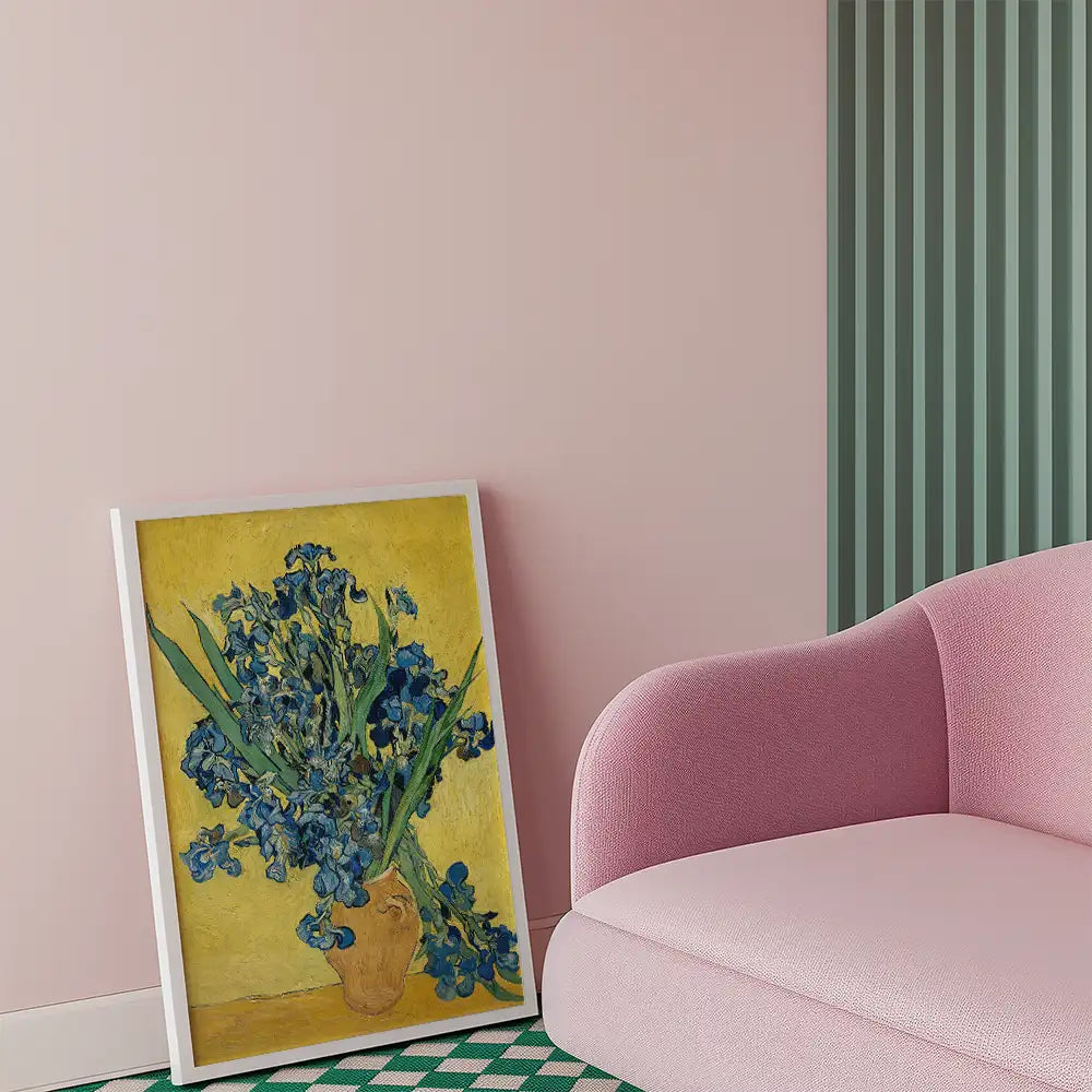 Framed painting of blue irises in a vase against a yellow background, displayed in a cozy interior with a pink sofa.