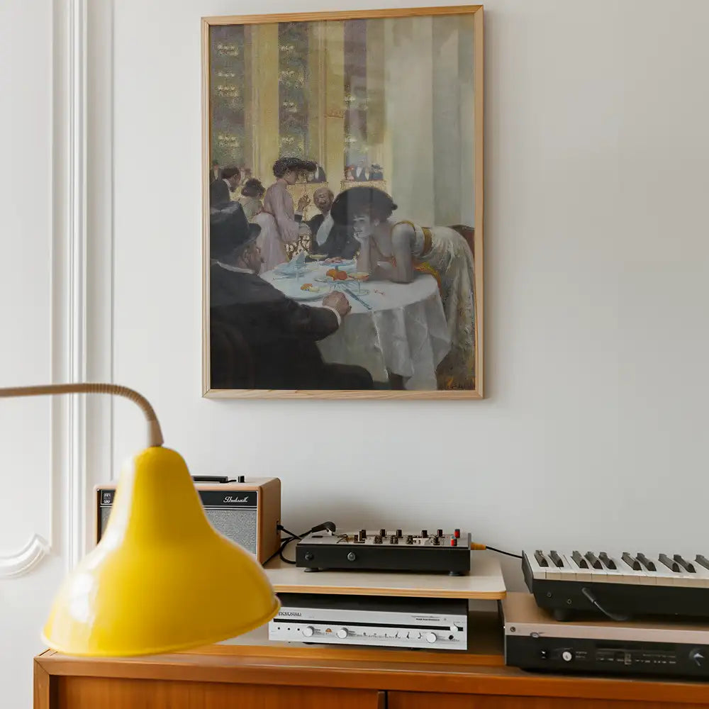 Framed painting of a social gathering at a dining table, featuring diverse figures in elegant attire, displayed on a white wall above a wooden sideboard with decorative objects.