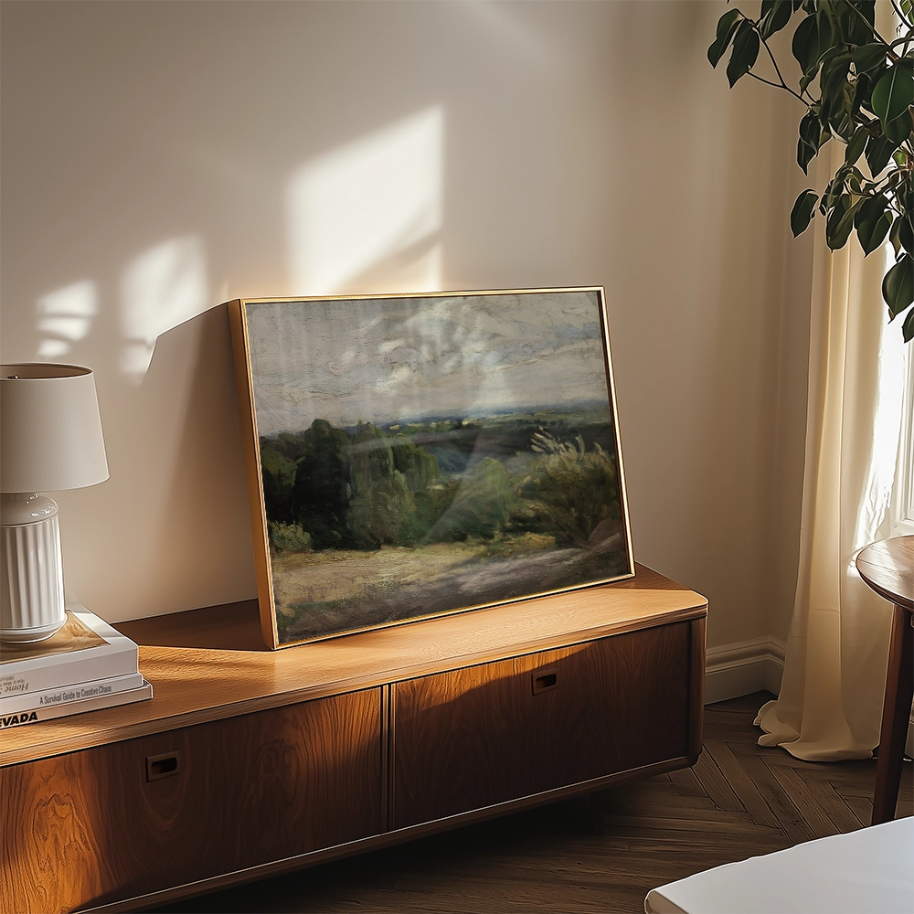 Framed landscape painting displayed on a wooden console table beside a lamp and potted plant, with natural sunlight illuminating the scene.