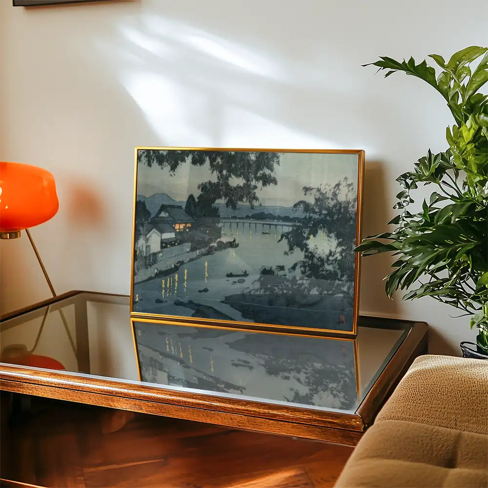 Framed painting of a serene riverside scene with reflections, displayed on a wooden table beside a plant and an orange lamp.
