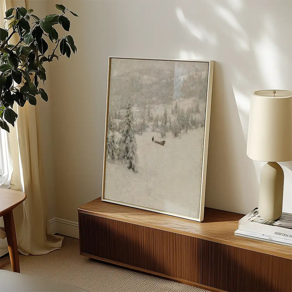 Framed landscape painting of a snowy scene with trees and a distant figure, displayed on a wooden console table next to a lamp and plant.