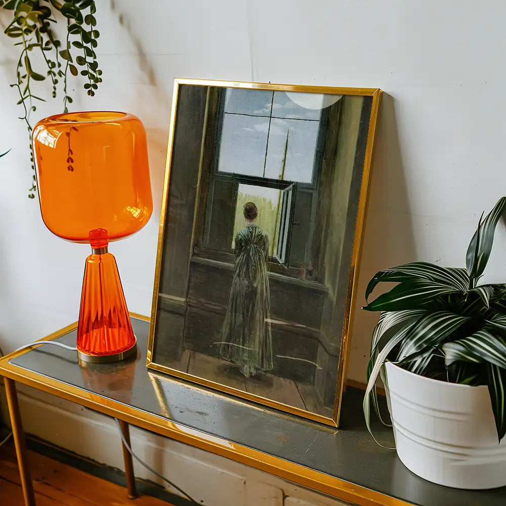 Painting of a woman looking out a window, displayed in a gold-framed picture on a table alongside an orange lamp and a houseplant.