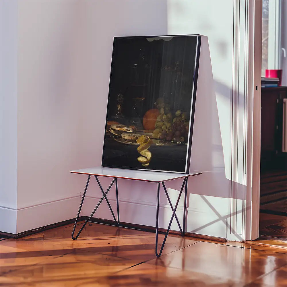 A framed painting featuring a still life composition with fruit and snacks displayed on a table, leaning against a wall in a sunlit room.