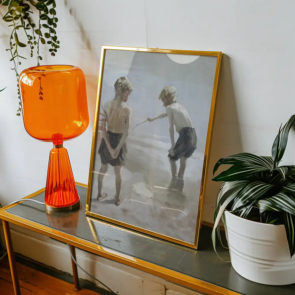 Framed painting of two children playing by the water, displayed on a table alongside an orange lamp and a potted plant.